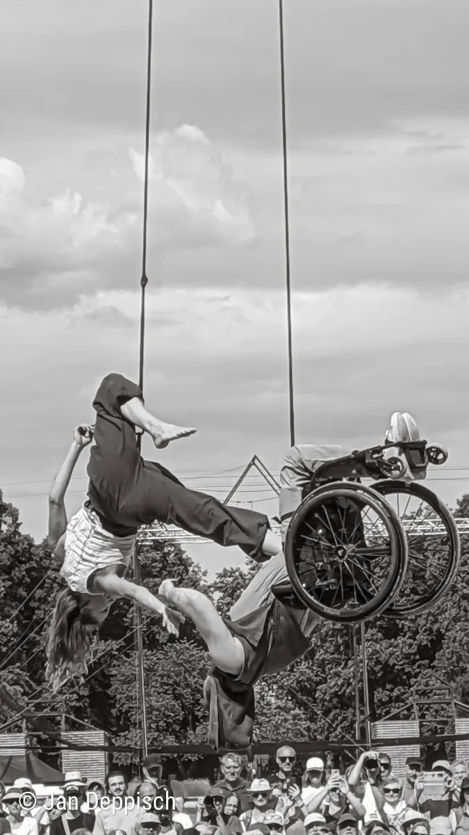 Two aerial performers mid-swing on ropes high above an outdoor festival crowd; one is a wheelchair user upside-down in mid-air, wheels above head. Black-and-white photo. Copyright: Jan Deppisch
